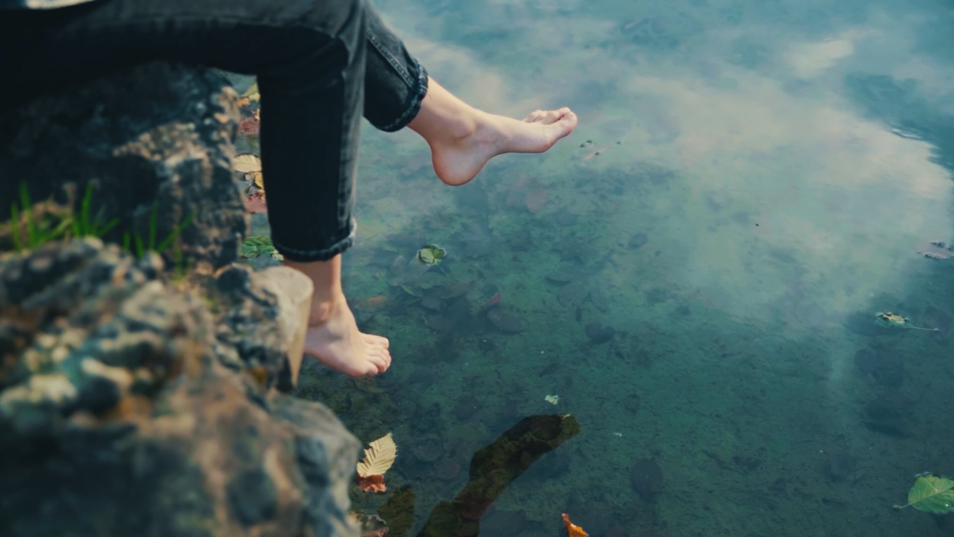 The legs of a female sitting on the border of a pond, swinging her legs back and forth above the water