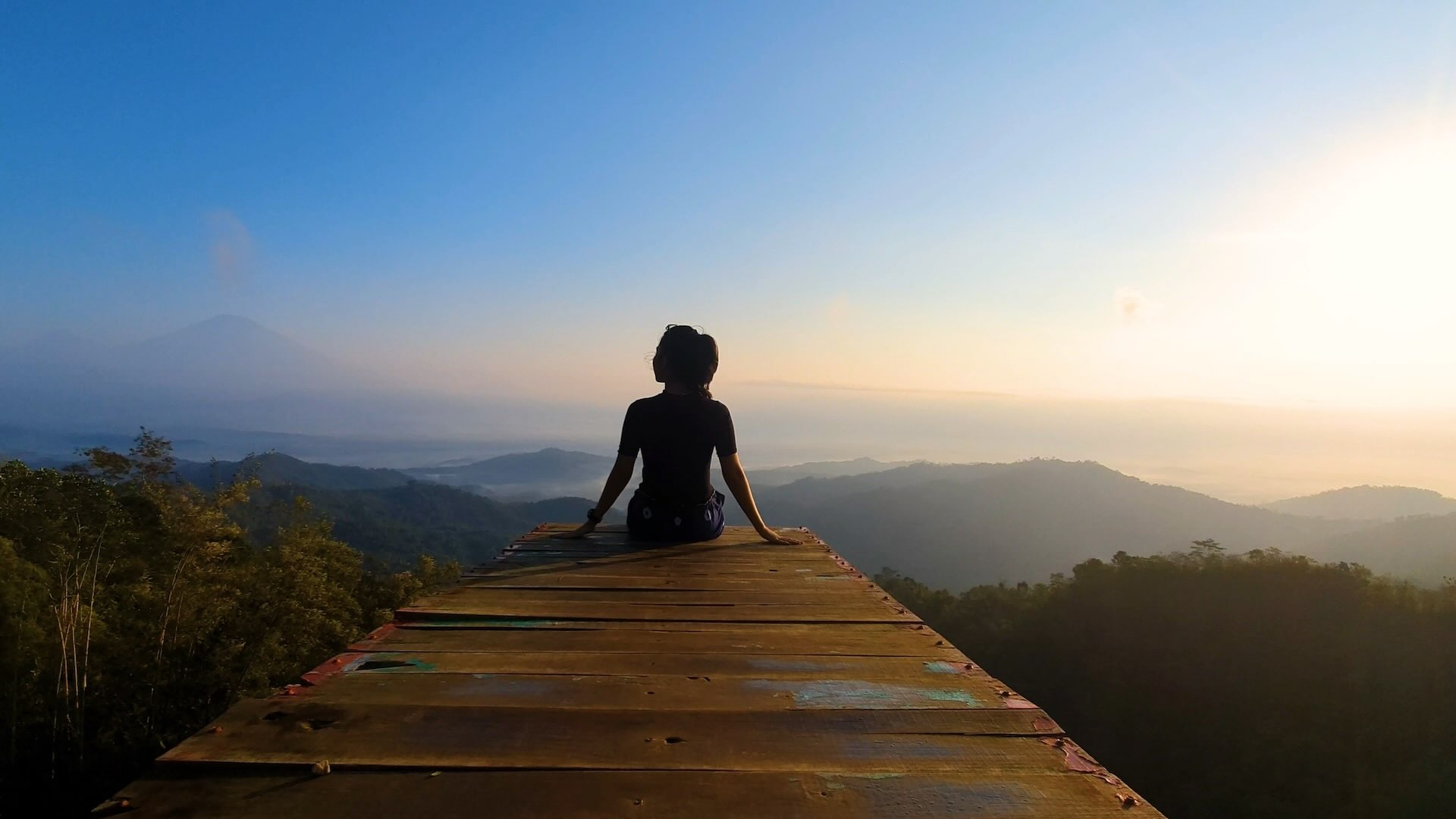 A woman viewed from the back, sitting on the border of a wooden terrace, on the top of a mountain, admiring the view