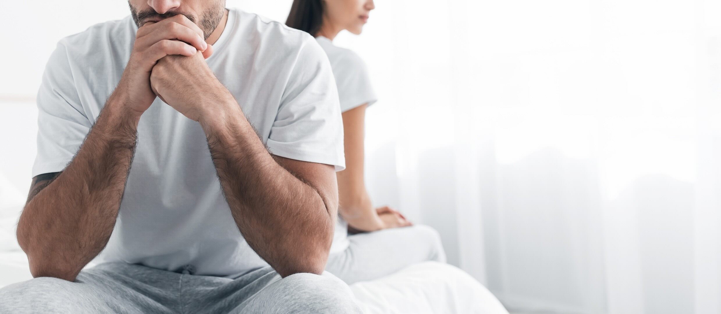 카메라를 향해 앉아 있는 백인 남성과 그 뒤에 있는 백인 여성이 침대 가장자리에 등을 맞대고 앉아 있는 것처럼 보이는 긴장된 상황