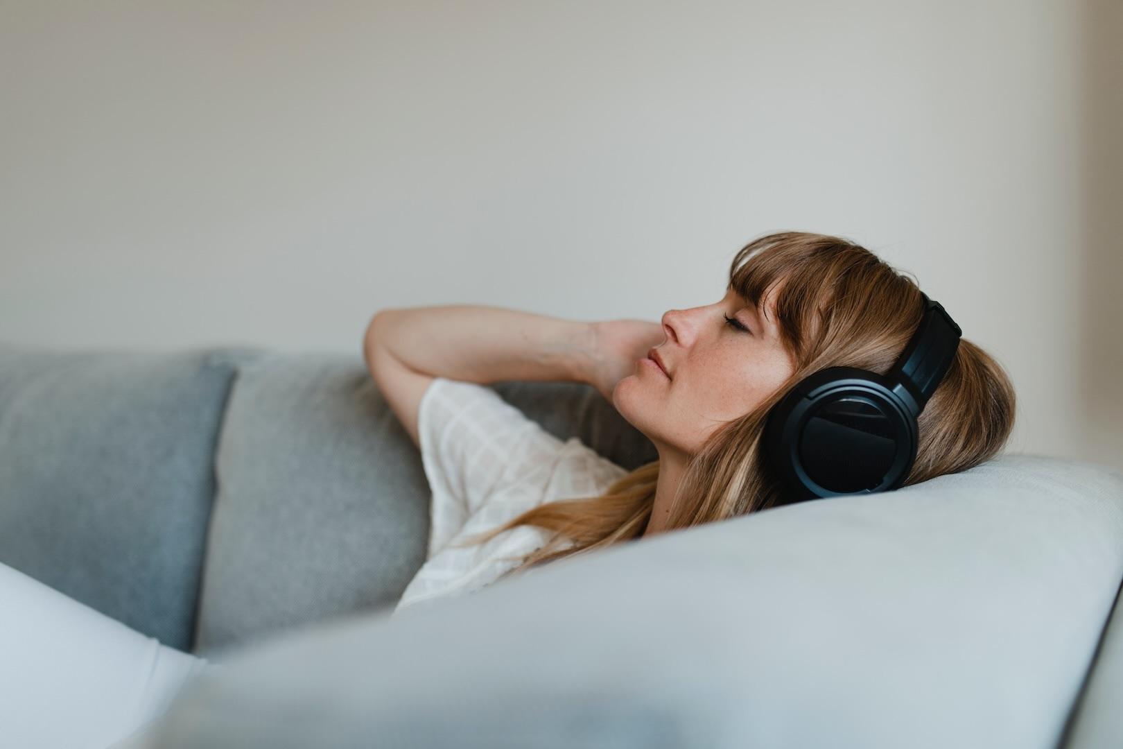 Femme blanche allongée sur un canapé avec un casque audio noir et les yeux fermés, se relaxant, avec une main sur le casque