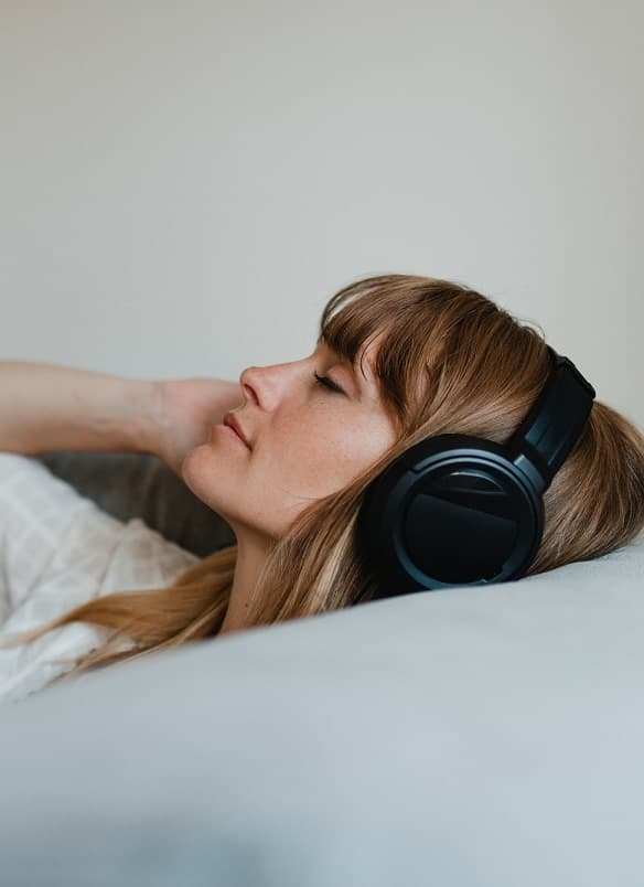 White female laying down on a couch with black headphones, and eyes closed, relaxing, with one hand on the headphones
