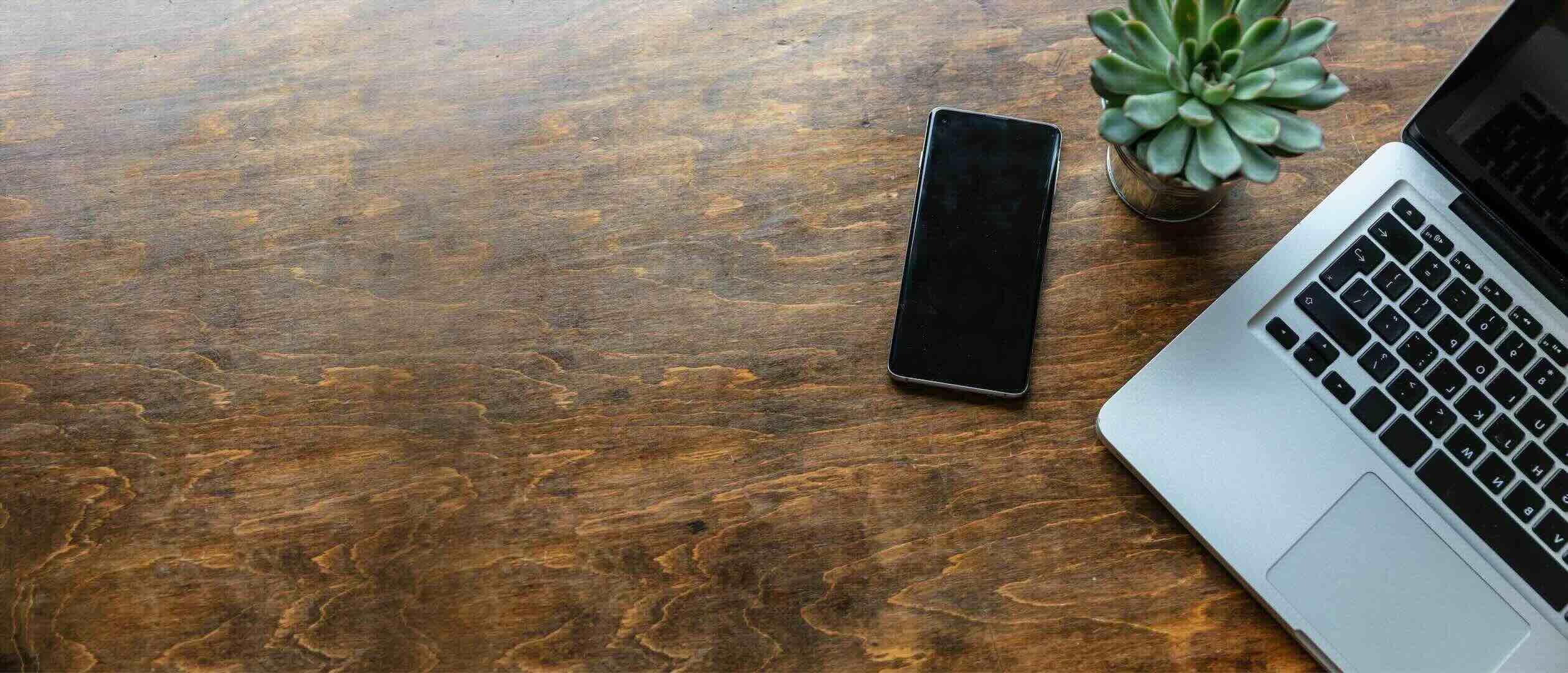 Desk made of wood with a laptop, a phone, and a plant on top of it, viewed from above