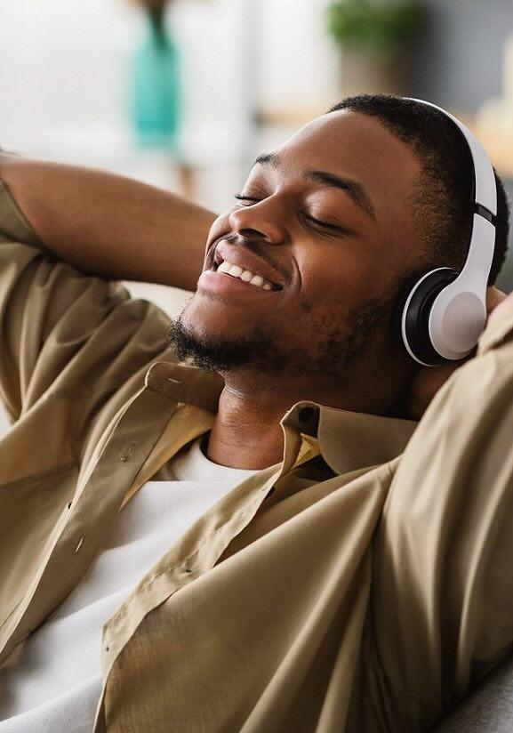 Homme noir portant un casque audio avec les yeux fermés, détendu sur un canapé, avec les mains derrière la tête