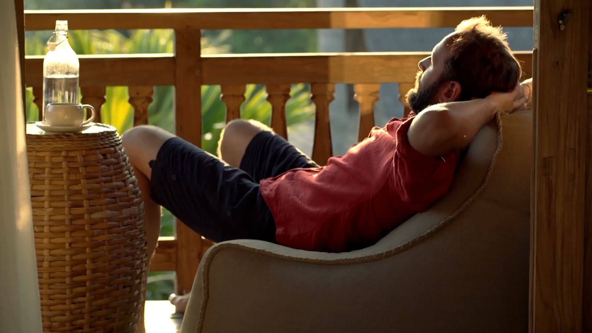 Un homme allongé sur une chaise longue sur une terrasse avec la nature en arrière-plan, se relaxant et mettant un chapeau sur son visage, pour se détendre